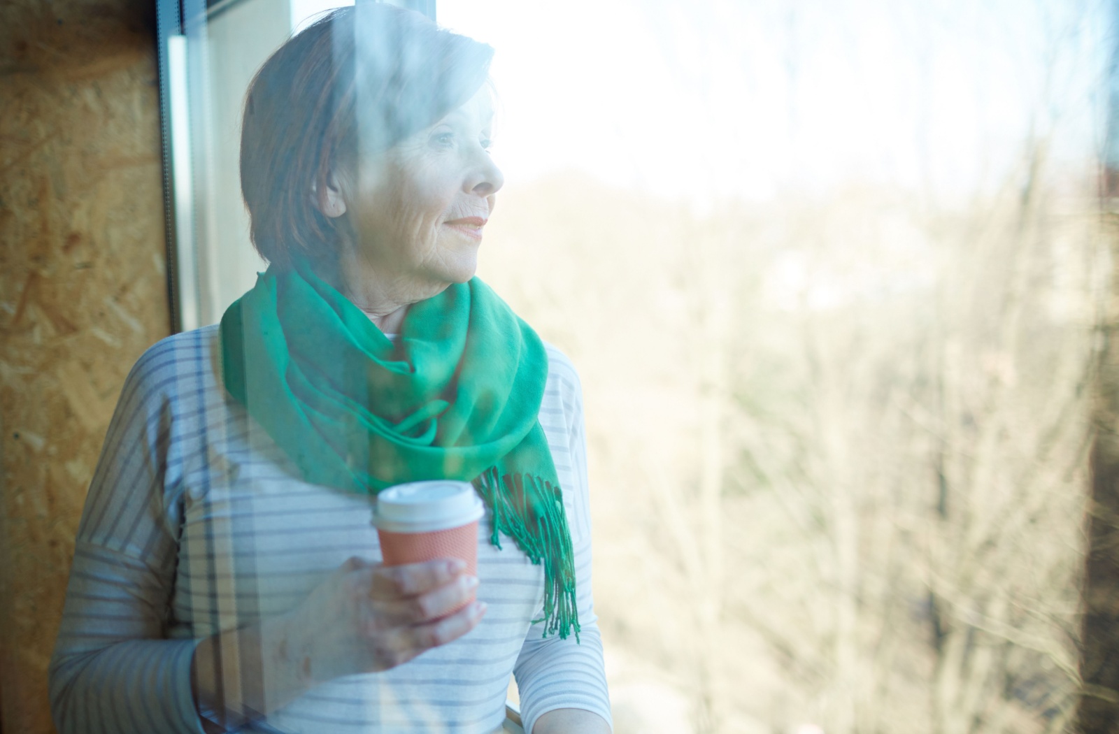 An older adult in a green scarf holds a coffee and looks out the window in late fall to look for inspiration for their next art project