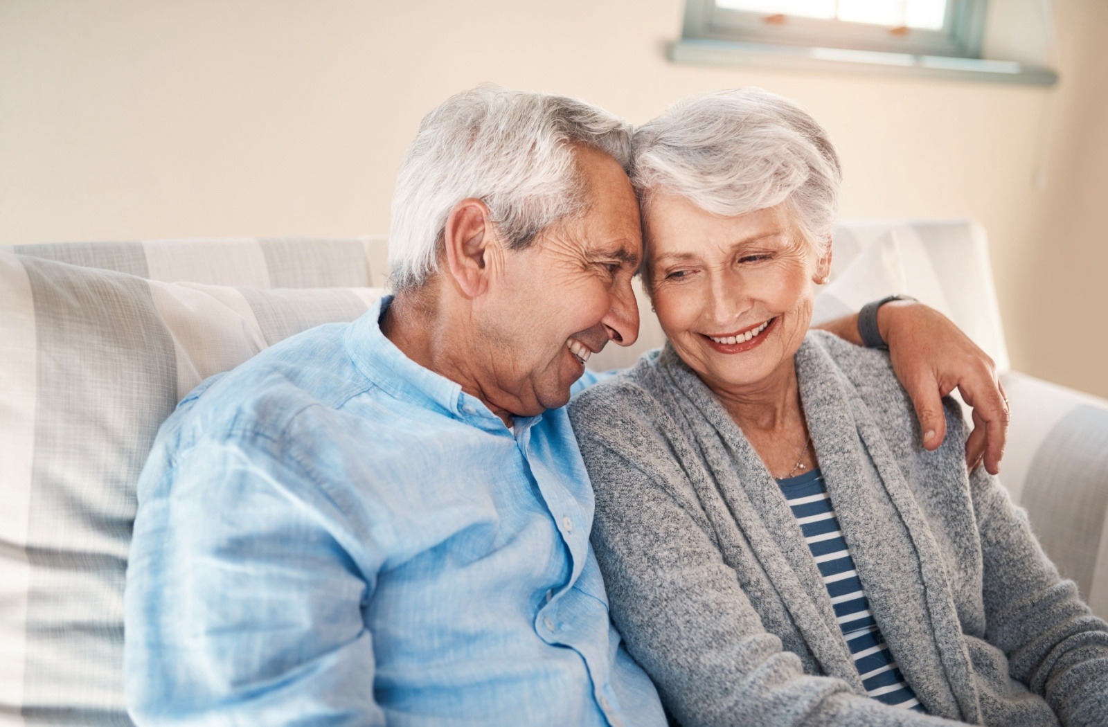 A senior couple in the process of downsizing embraces while sitting on a striped gray couch.
