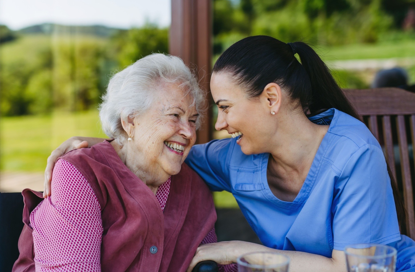 A caregiver laughs with a senior living resident while sitting at a table outside.
